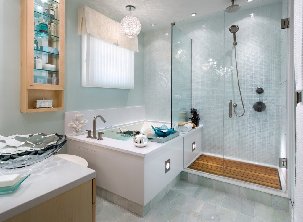 Updated bathroom design with light blue walls and decor, glass shower, round chandelier ball above the tub area, glass sink bowl and tiled floor.